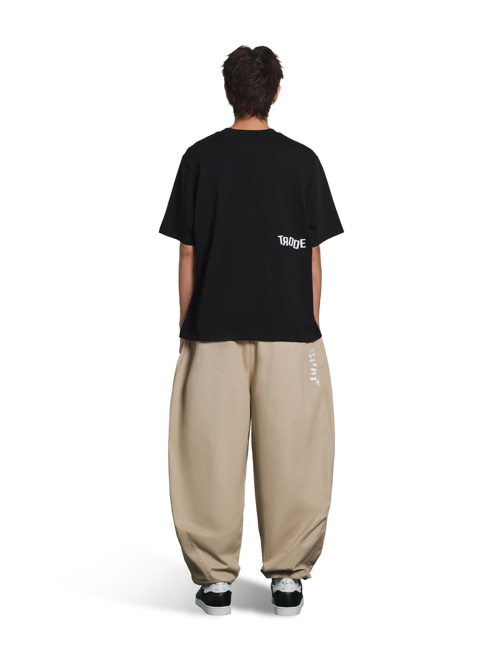 A model shown from the back wearing a black T-shirt with a small white 'TOKYO' graphic on the left shoulder, paired with baggy beige pants and black sneakers, against a plain white background.