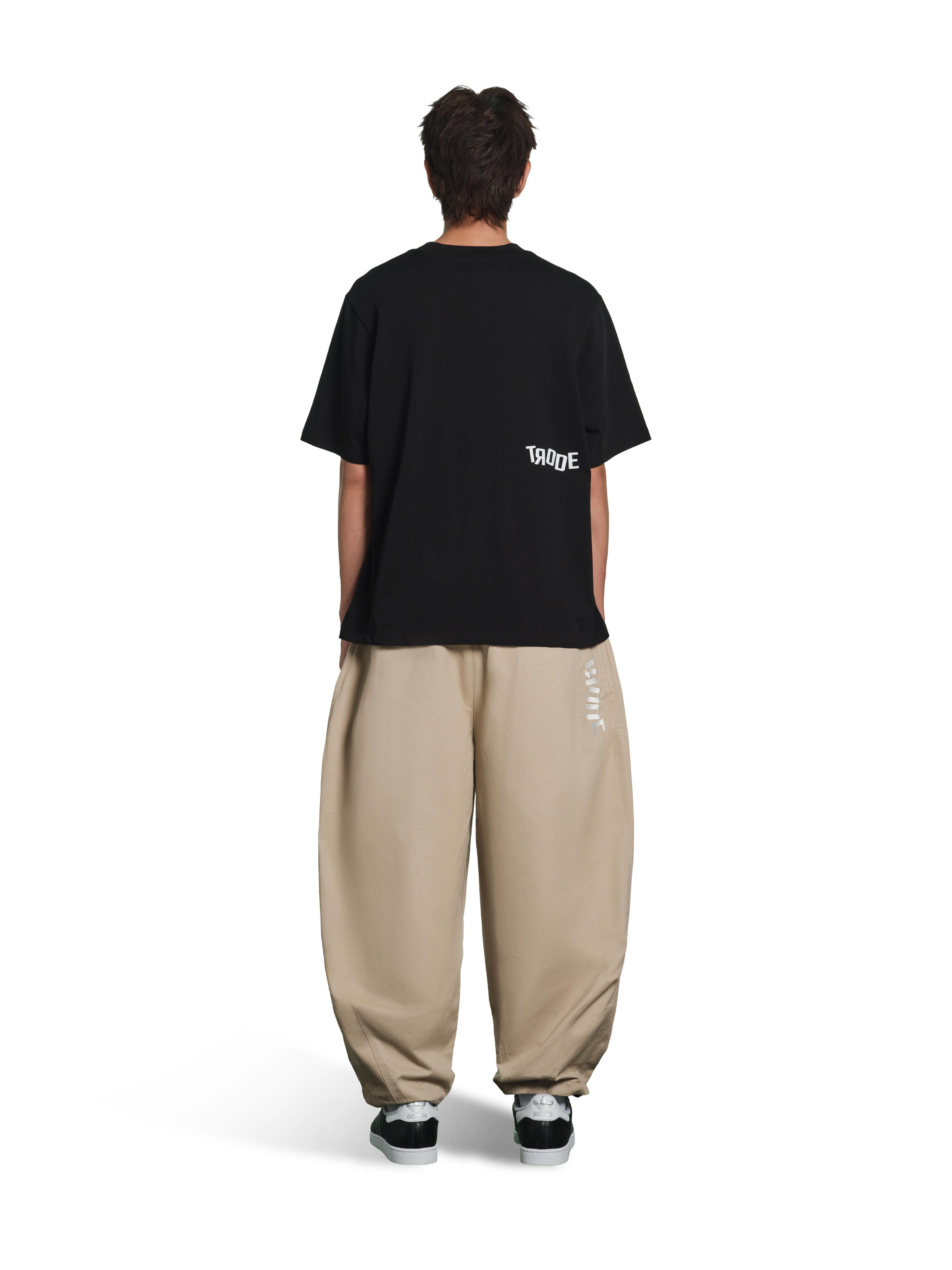 A model shown from the back wearing a black T-shirt with a small white 'TOKYO' graphic on the left shoulder, paired with baggy beige pants and black sneakers, against a plain white background.