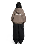 A model shown from the back wearing a taupe-brown cropped hoodie with 'TRODE' printed in white across the back, paired with black baggy sweatpants featuring vertical white 'TRODE' branding on the side.