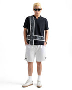A man modeling a black short-sleeve polo shirt with a bold white and gray cross design on the front, paired with white shorts, white socks, and white sneakers, set against a plain white background.