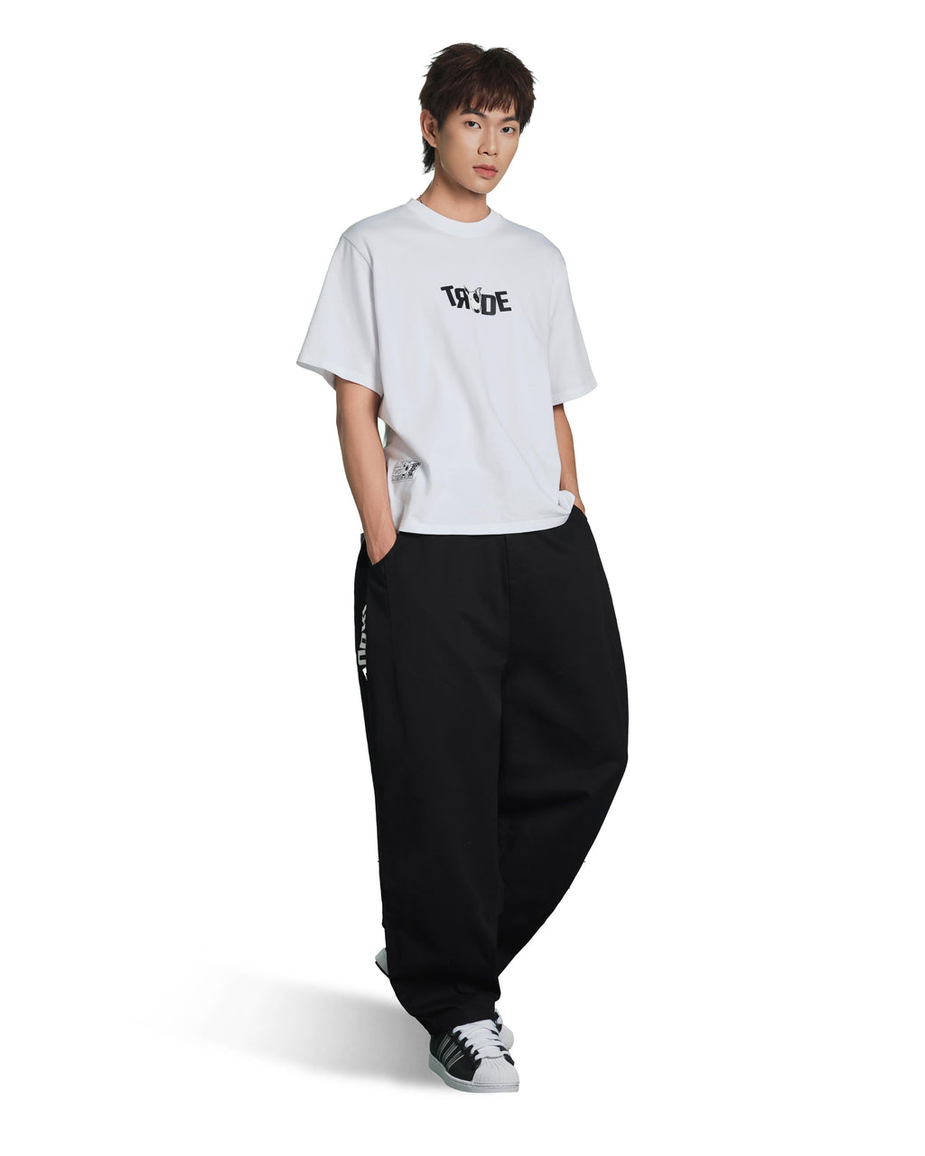 A model wearing a white short-sleeve t-shirt with a black graphic print on the chest, paired with loose black sweatpants and black-and-white sneakers, set against a plain white background.