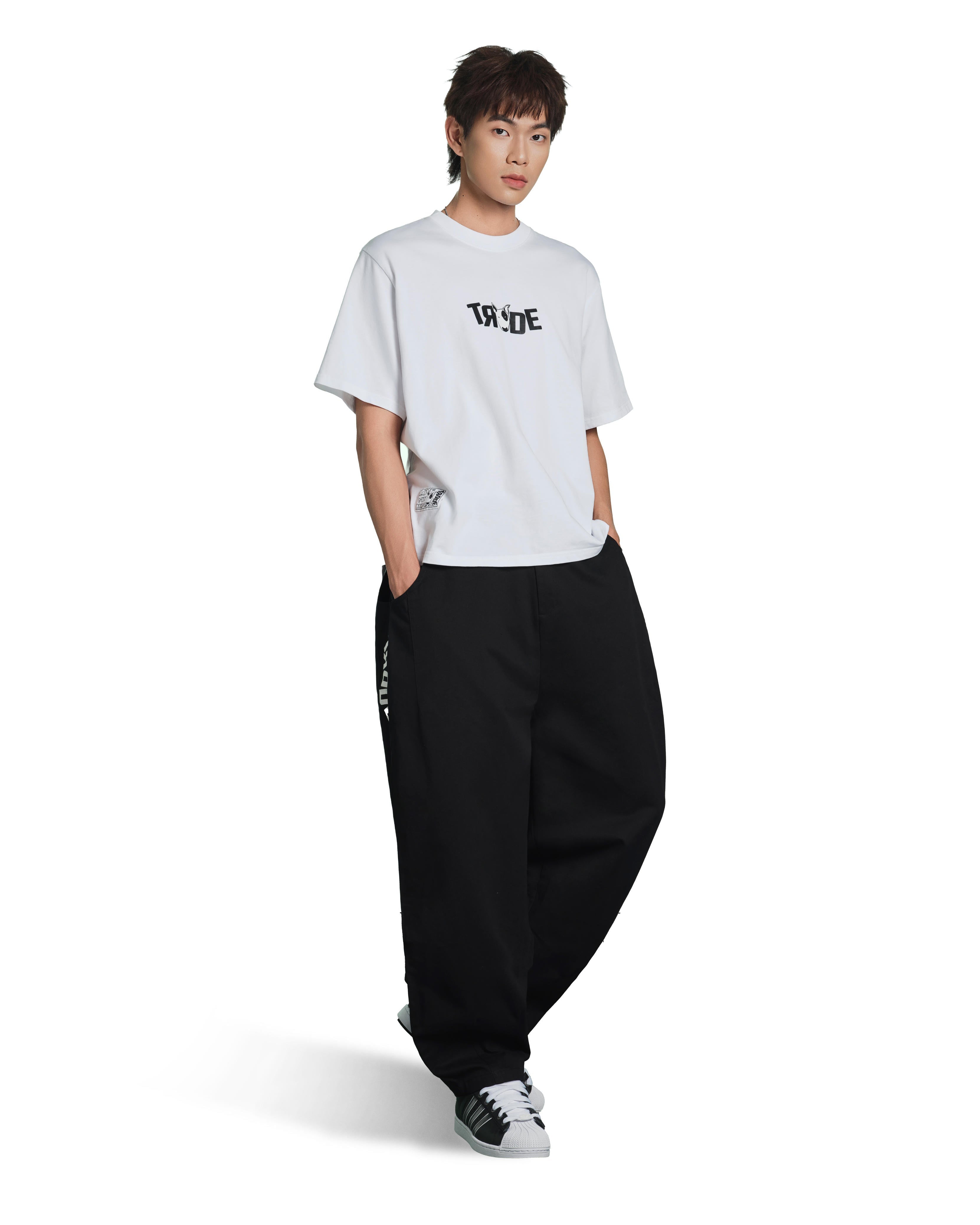 A model wearing a white short-sleeve t-shirt with a black graphic print on the chest, paired with loose black sweatpants and black-and-white sneakers, set against a plain white background.