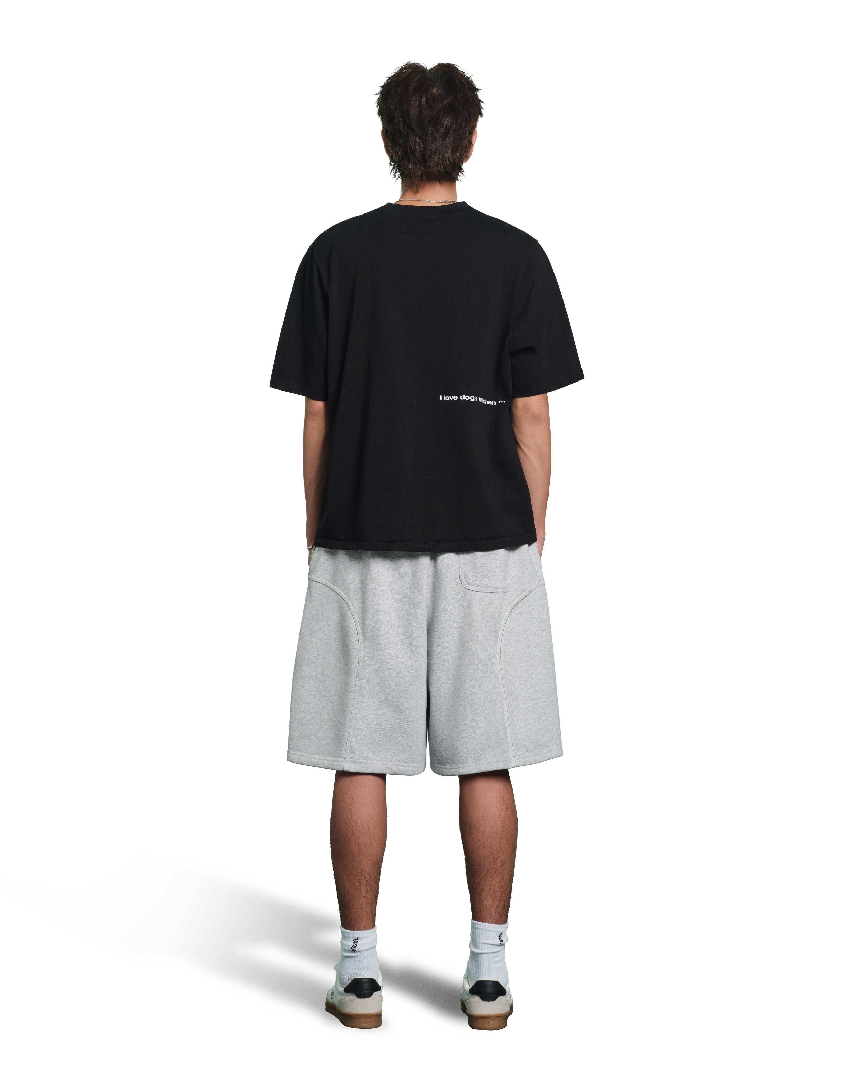 A model shown from the back wearing an oversized black t-shirt with small white text on the back, light gray heathered shorts, white socks, and sneakers, against a plain white background.