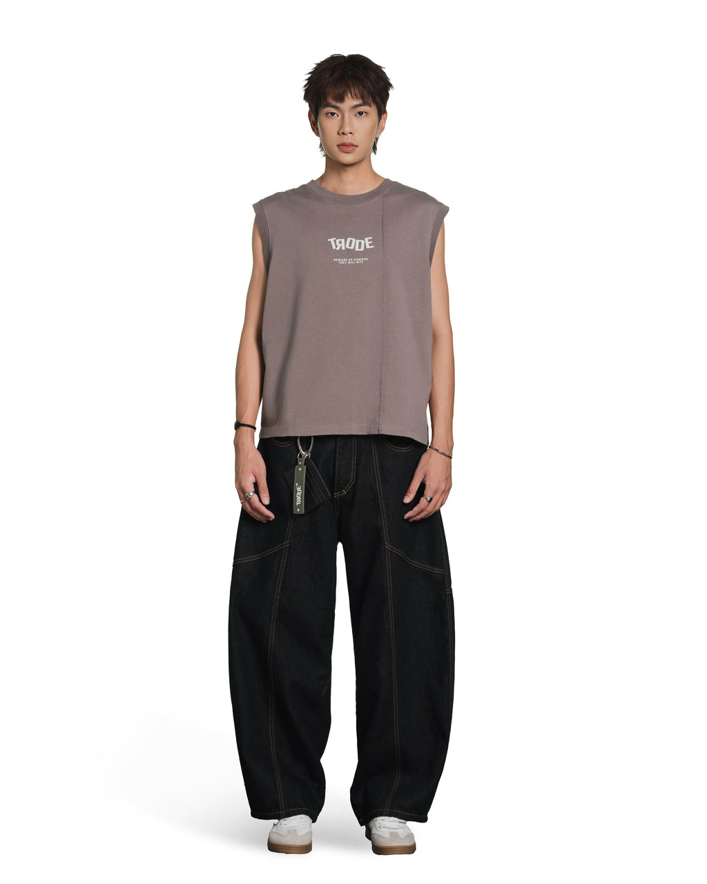 A male model stands against a plain white studio background wearing a sleeveless taupe-brown t-shirt with a white graphic print on the chest, paired with loose black pants and white sneakers.