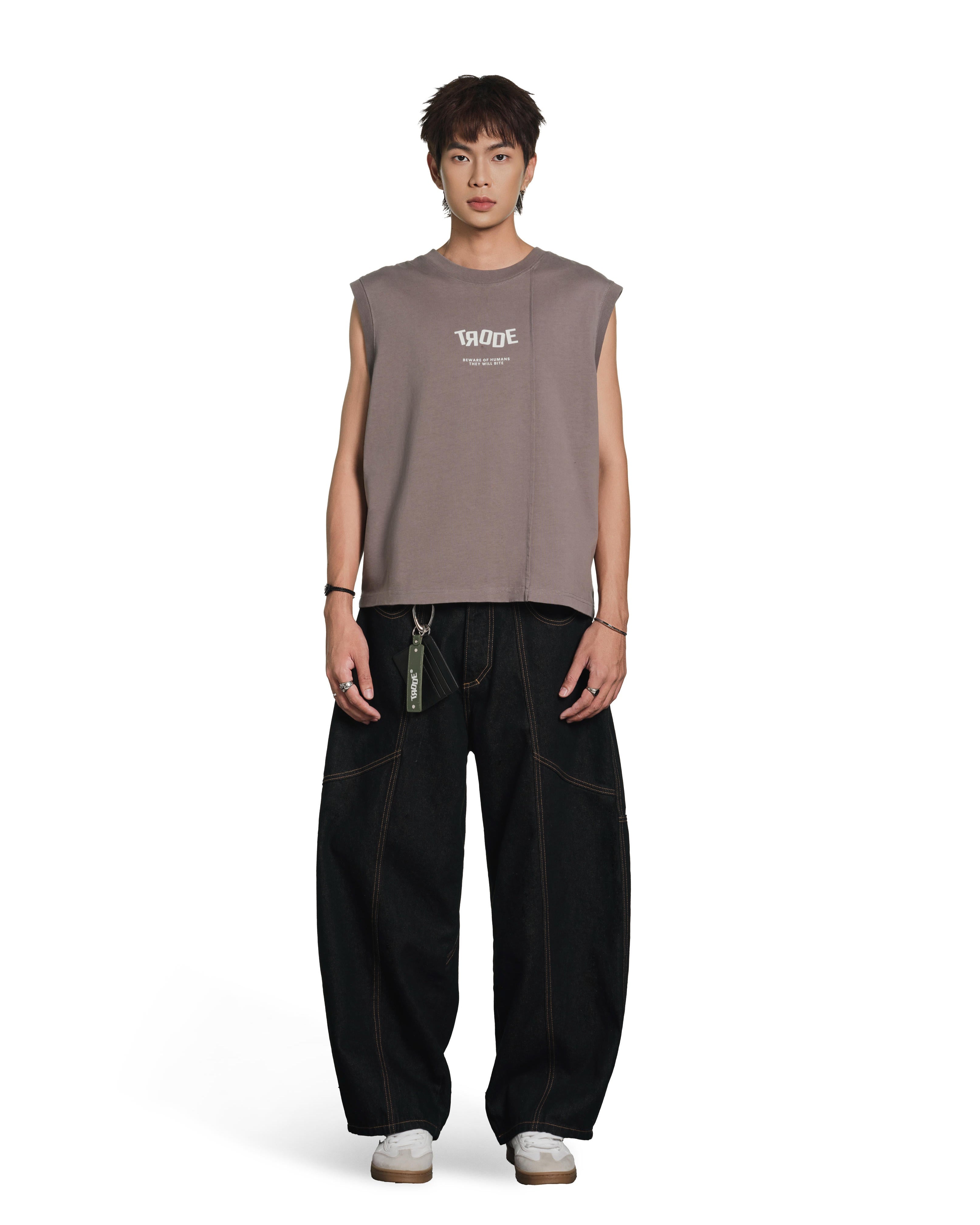 A male model stands against a plain white studio background wearing a sleeveless taupe-brown t-shirt with a white graphic print on the chest, paired with loose black pants and white sneakers.