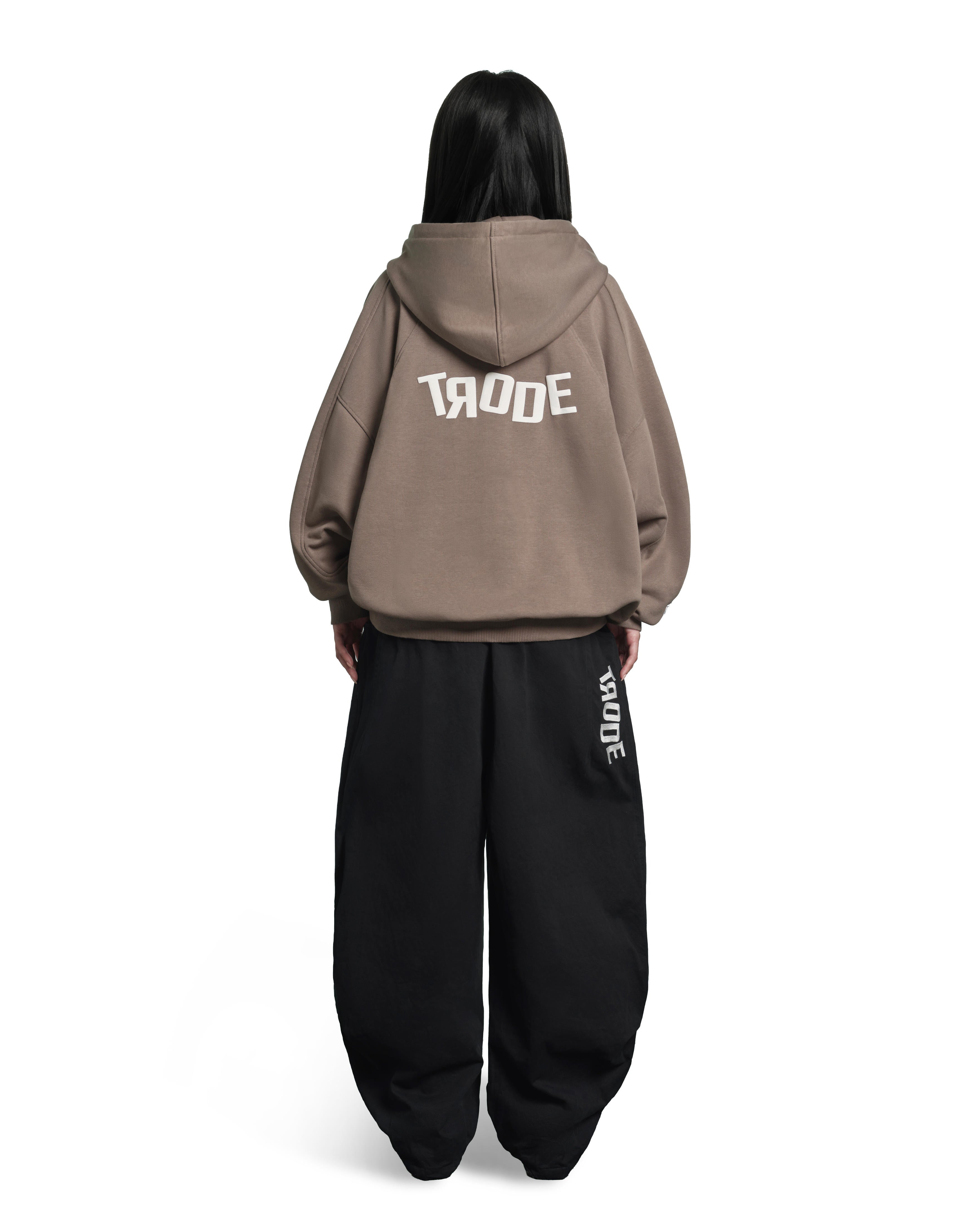 A model shown from the back wearing a taupe-brown cropped hoodie with 'TRODE' printed in white across the back, paired with black baggy sweatpants featuring vertical white 'TRODE' branding on the side.