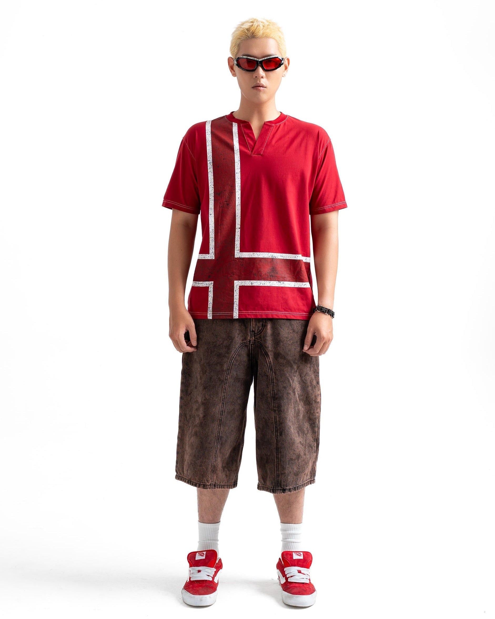 A model stands against a white studio background wearing a bright red short-sleeve t-shirt with white geometric block patterns, brown cropped shorts, red sneakers, white socks, and red sunglasses.