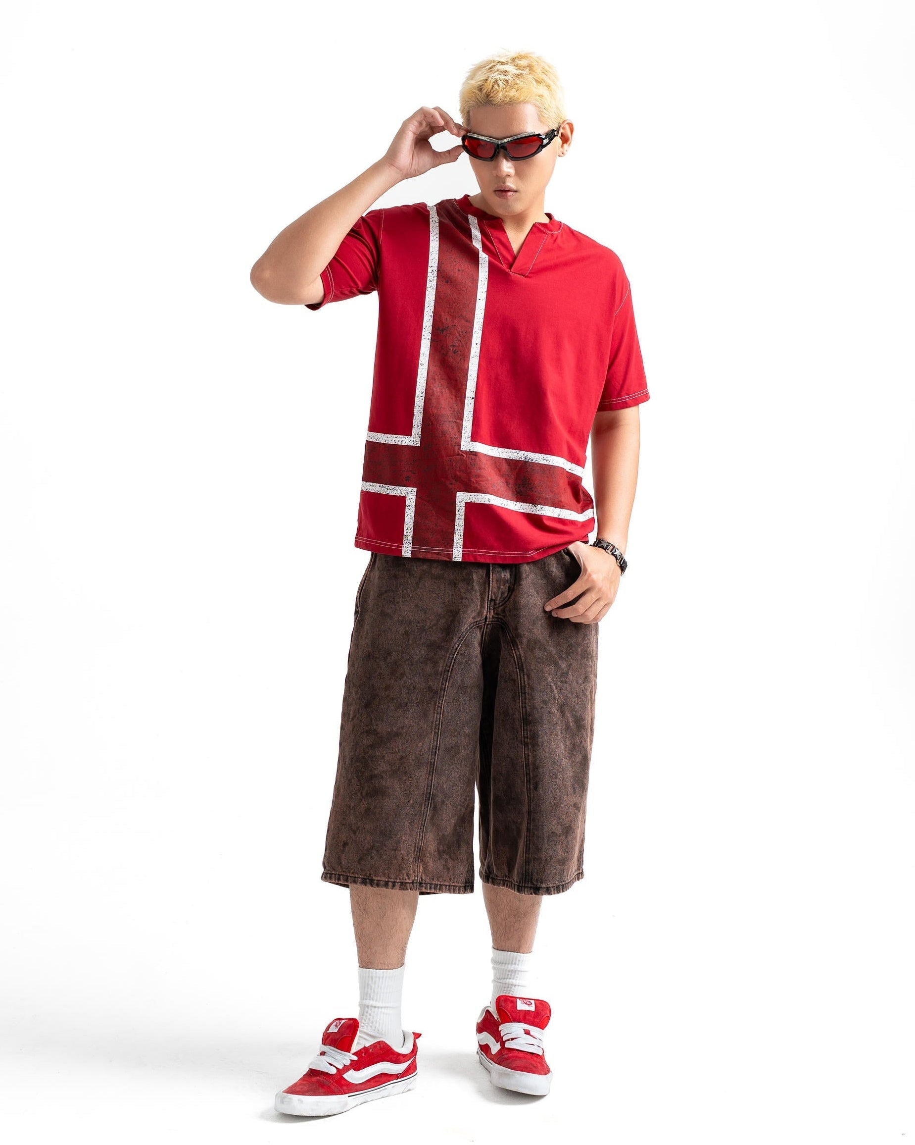 A full-body shot of a model wearing a red short-sleeve polo shirt with a white Nordic flag design, brown textured knee-length shorts, red and white sneakers, and red sunglasses, set against a plain white background.
