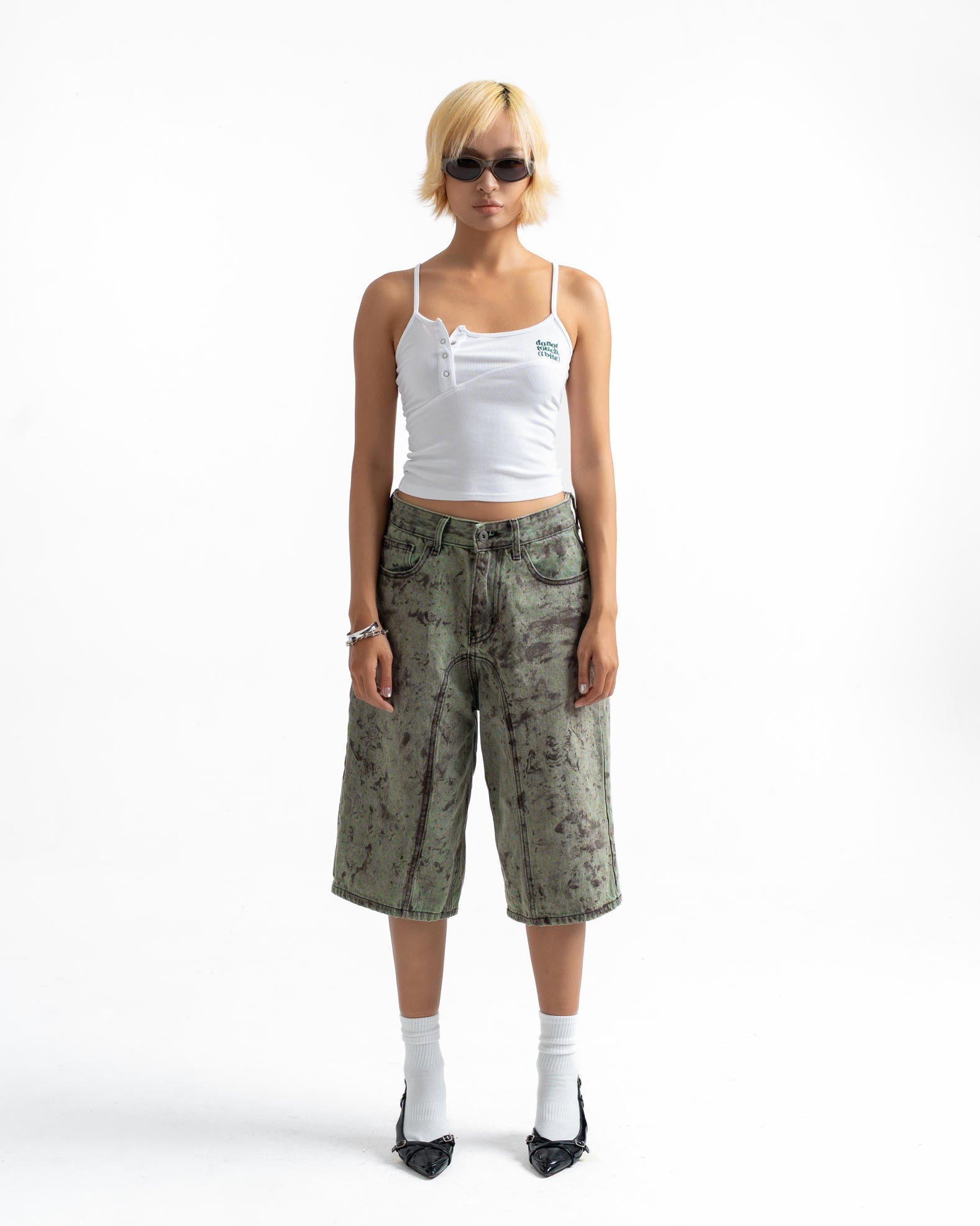 A model stands against a white studio background wearing a white cropped camisole, olive green camouflage cropped shorts, white socks, and black pointed shoes.