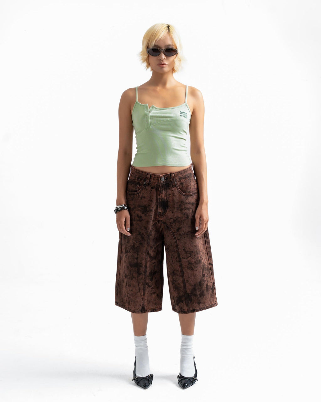 A model stands against a white studio background wearing a light green camisole, brown patterned cropped pants, white socks, and black shoes.