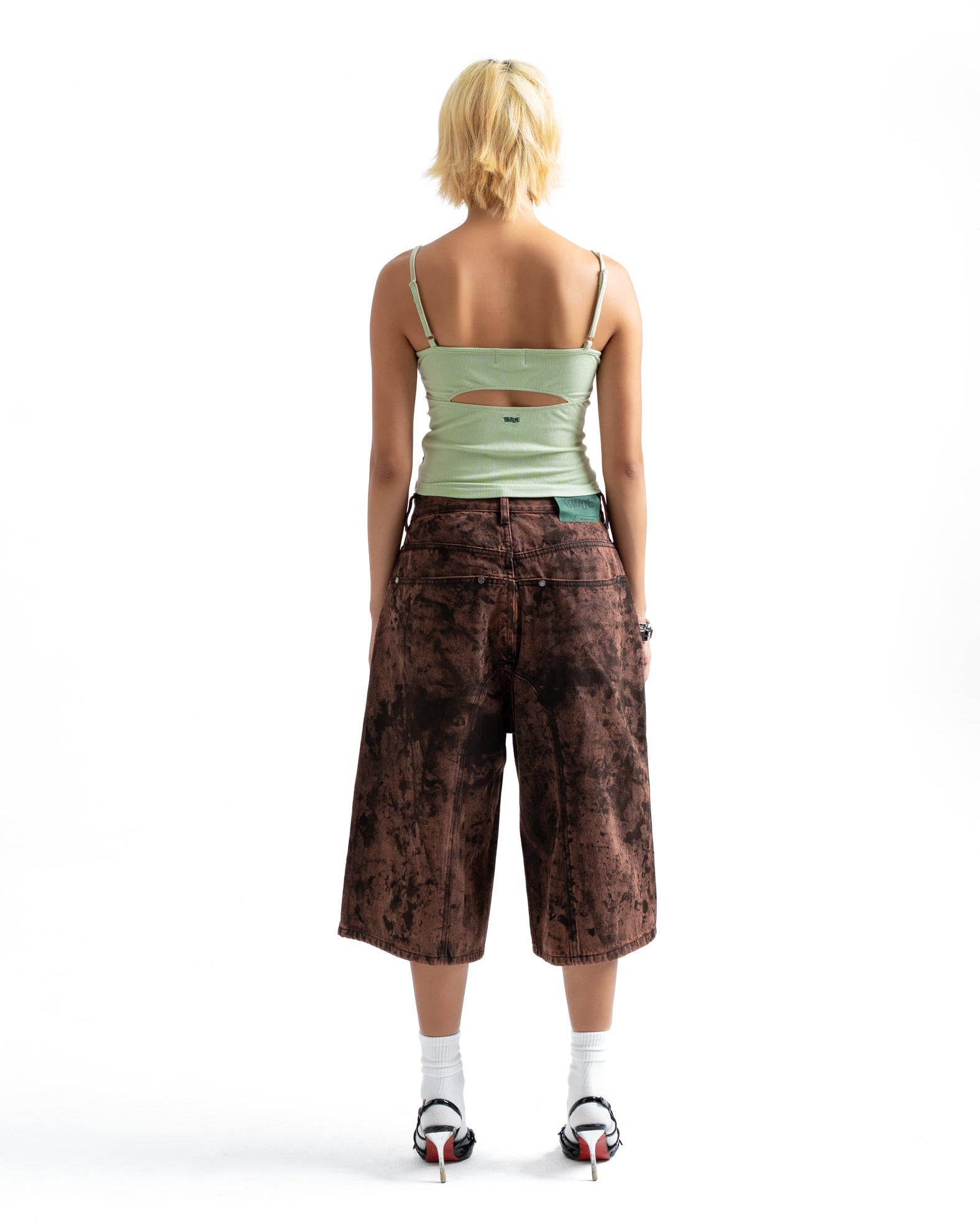 A model viewed from behind wearing a light green spaghetti-strap top with a cutout, brown cropped pants with a dark abstract pattern, white socks, and black high heels on a white studio background.