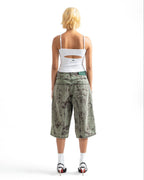 A model viewed from behind wearing a white cutout camisole, green camouflage cropped shorts, white socks, and black high heels on a white studio background.