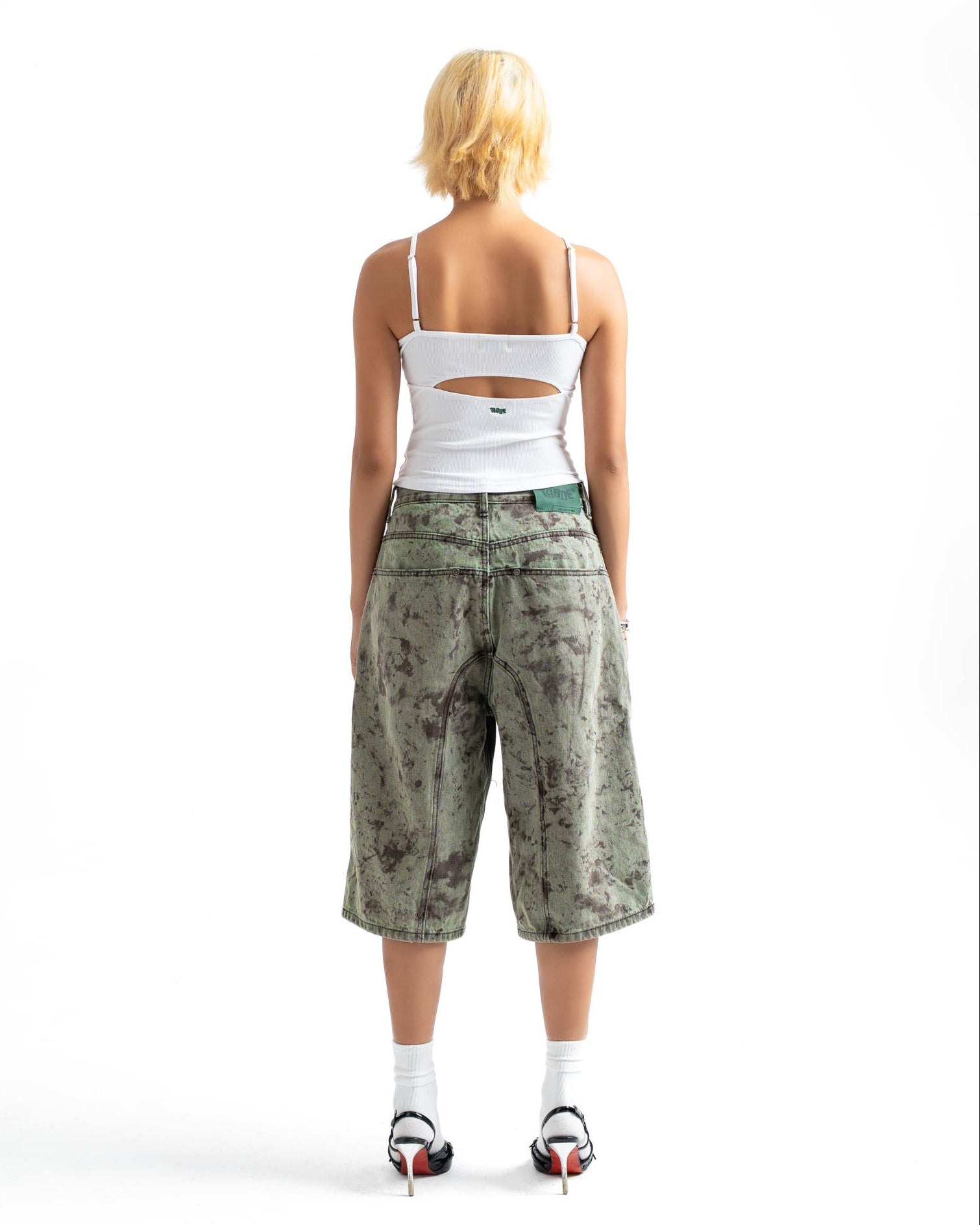A model viewed from behind wearing a white cutout camisole, green camouflage cropped shorts, white socks, and black high heels on a white studio background.