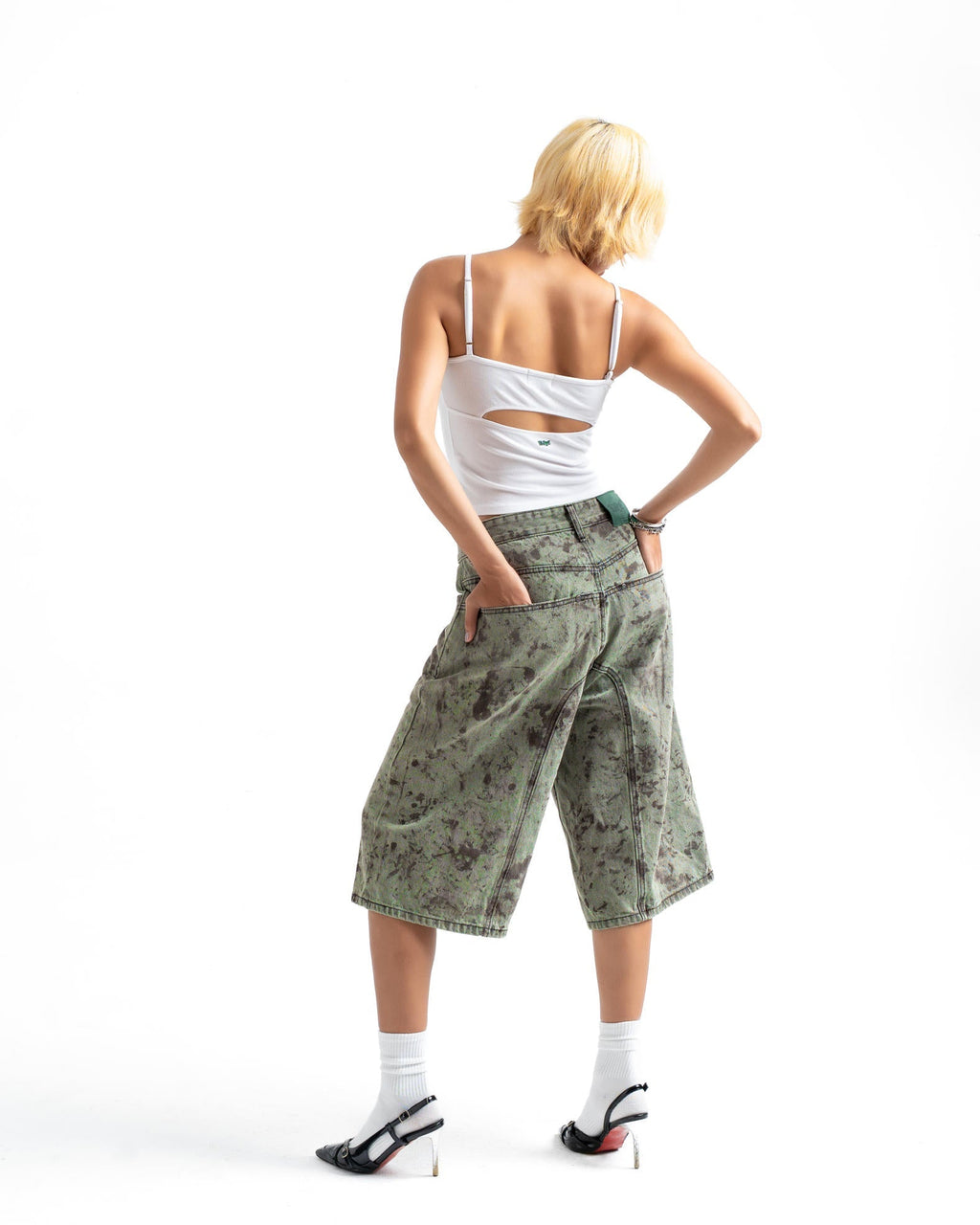 A model shown from behind wearing a white cutout camisole, green camouflage cropped shorts, white socks, and black high heels against a white background.