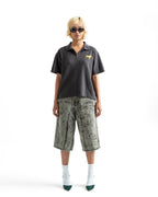 A model stands against a white studio background wearing a dark charcoal gray short-sleeve polo shirt with a small yellow embroidered logo on the left chest, paired with olive green and gray camouflage knee-length shorts, white socks, and dark green pointed shoes.