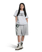 A model stands against a white background wearing a white graphic t-shirt with black text, light gray drawstring shorts, white socks, and off-white lace-up boots. The t-shirt features the text 'DEWARF ALL HUMANS' with a small illustration.