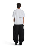 A model shown from the back wearing a white t-shirt with small black text on the back and black baggy sweatpants with a white vertical logo on the left leg, set against a plain white background.
